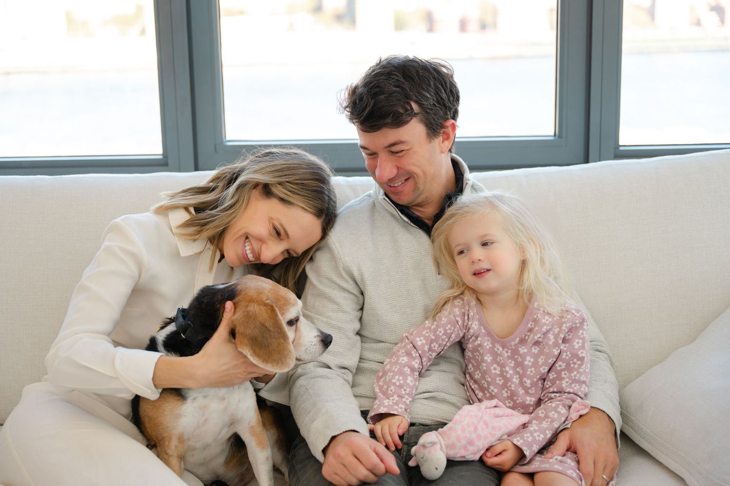 Happy family with a young girl and beagle dog sitting together on a white couch in a bright modern home.