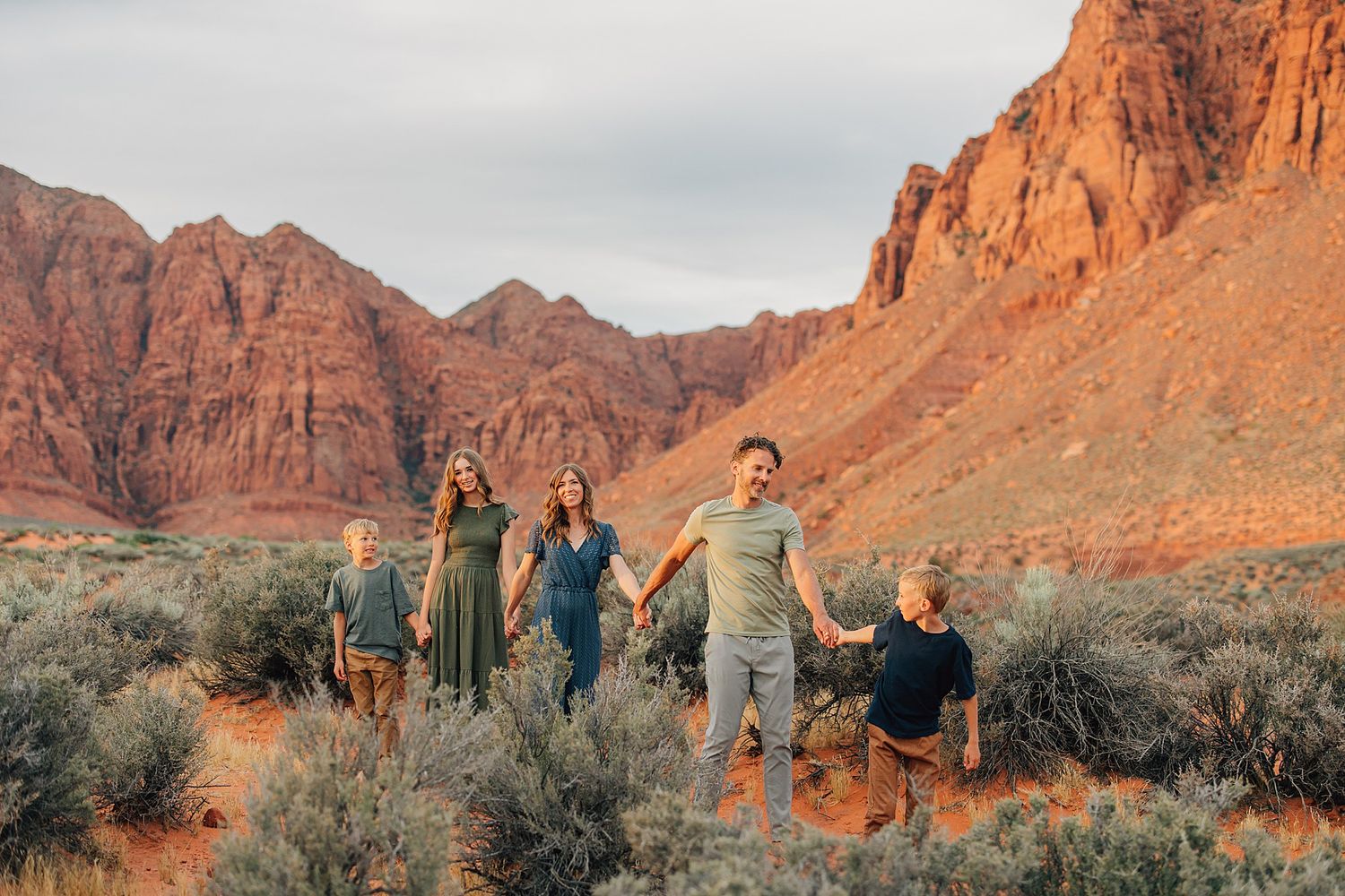 Family of five holding hands walking through red rock desert landscape with stunning sandstone cliffs backdrop.