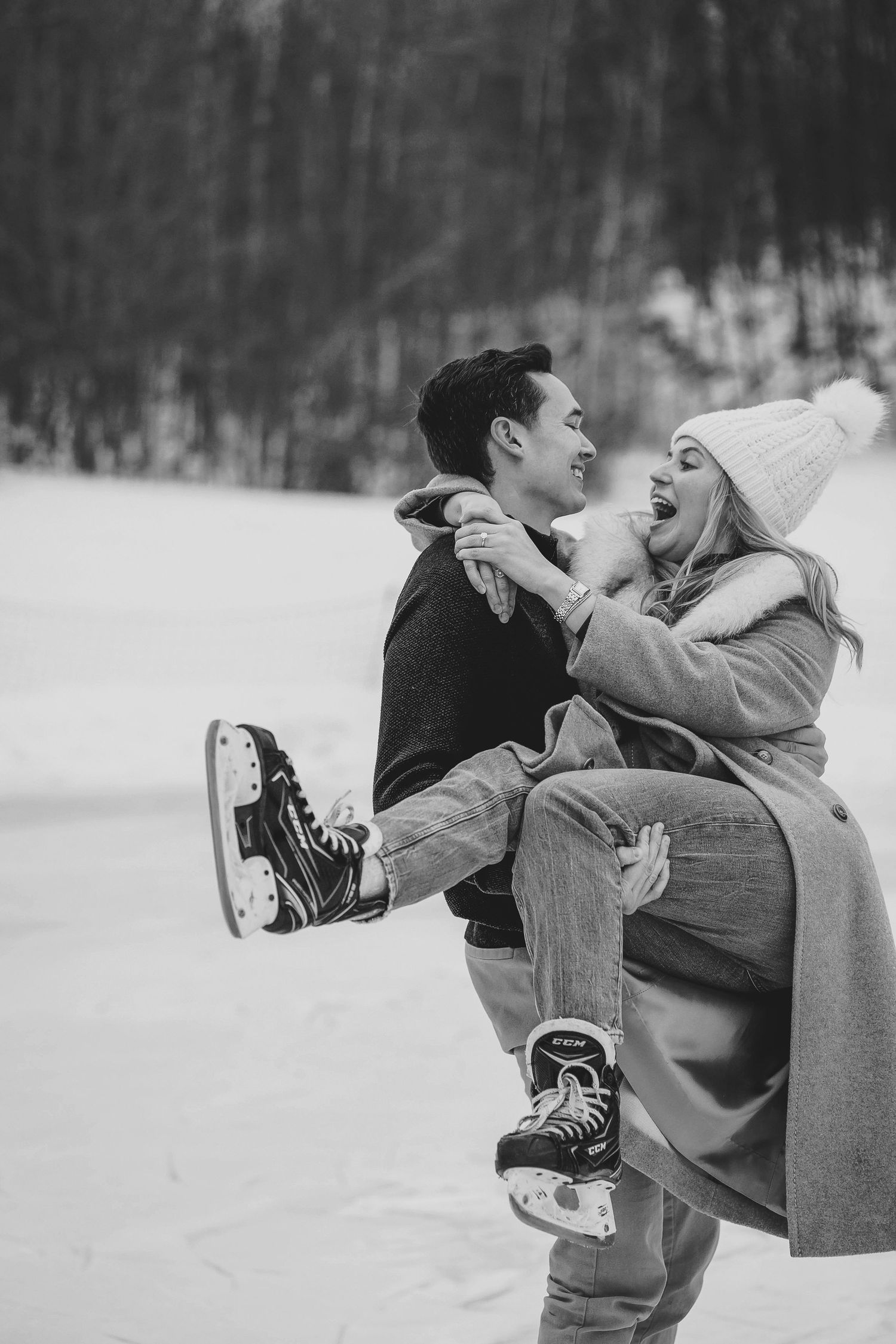 Black and white photo of a joyful couple ice skating in winter, man lifting woman in cozy hats and coats outdoors.