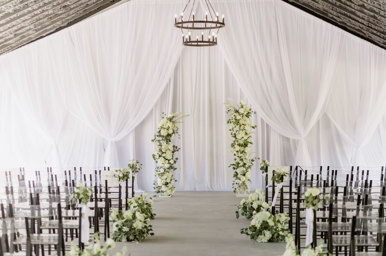 Bride and groom walk down the aisle at an elegant white-draped outdoor wedding ceremony with floral arrangements.