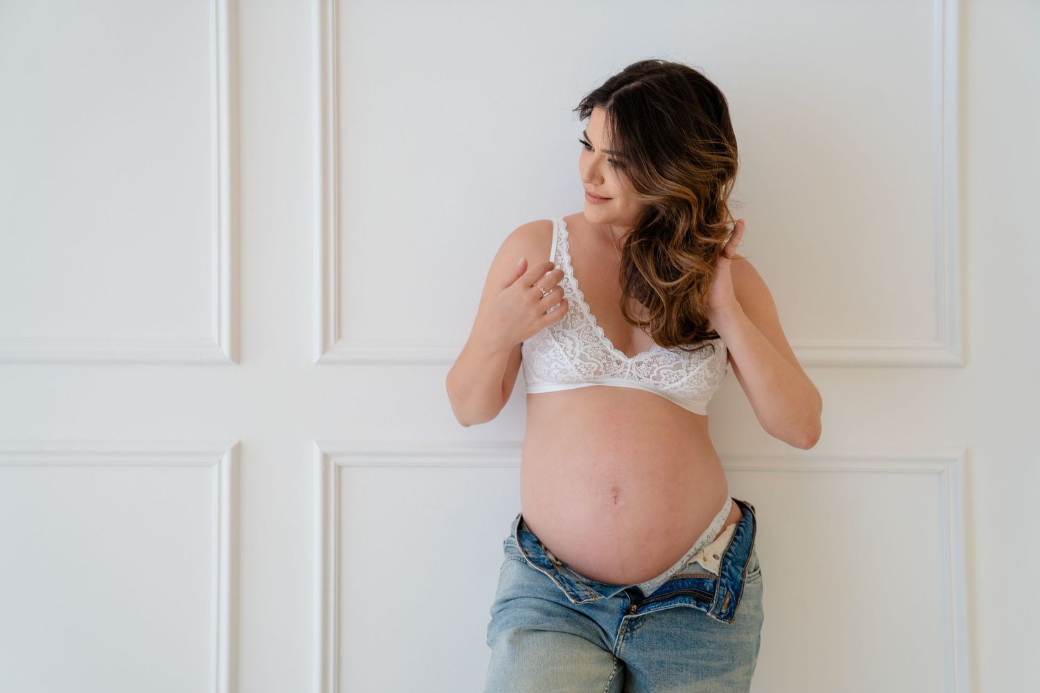 Pregnant woman in white lace bralette and jeans posing against white paneled wall, showcasing baby bump.
