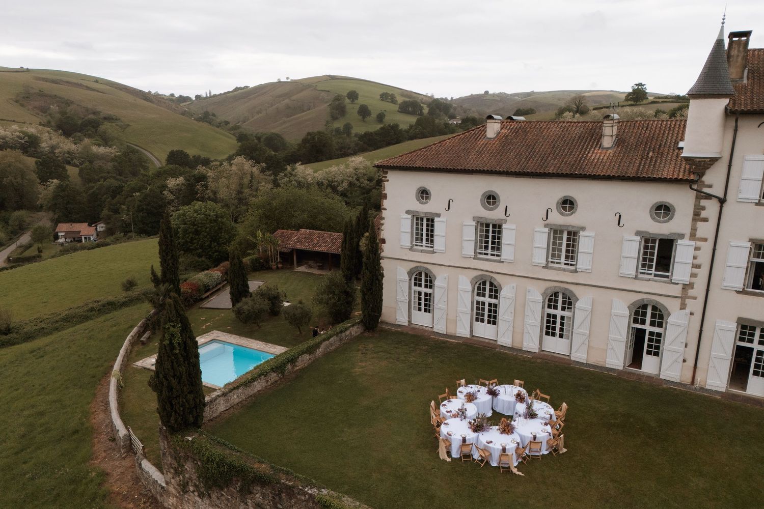 Aerial view of an elegant white French chateau with a pool, manicured lawns, and a formal outdoor dining setup in rolling hills.