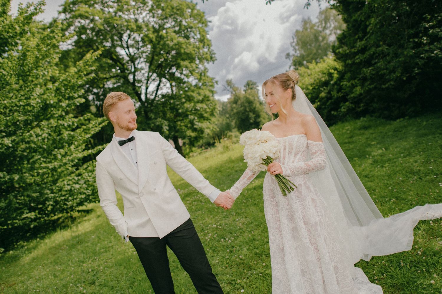 Newlywed couple holding hands in a lush green park, bride in lace gown with veil, groom in white tuxedo.