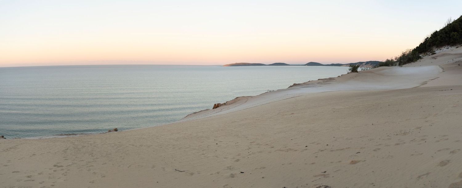 Serene Carlo Sandblow at dusk with calm waters, rolling dunes, and distant islands under a pastel sunset sky.