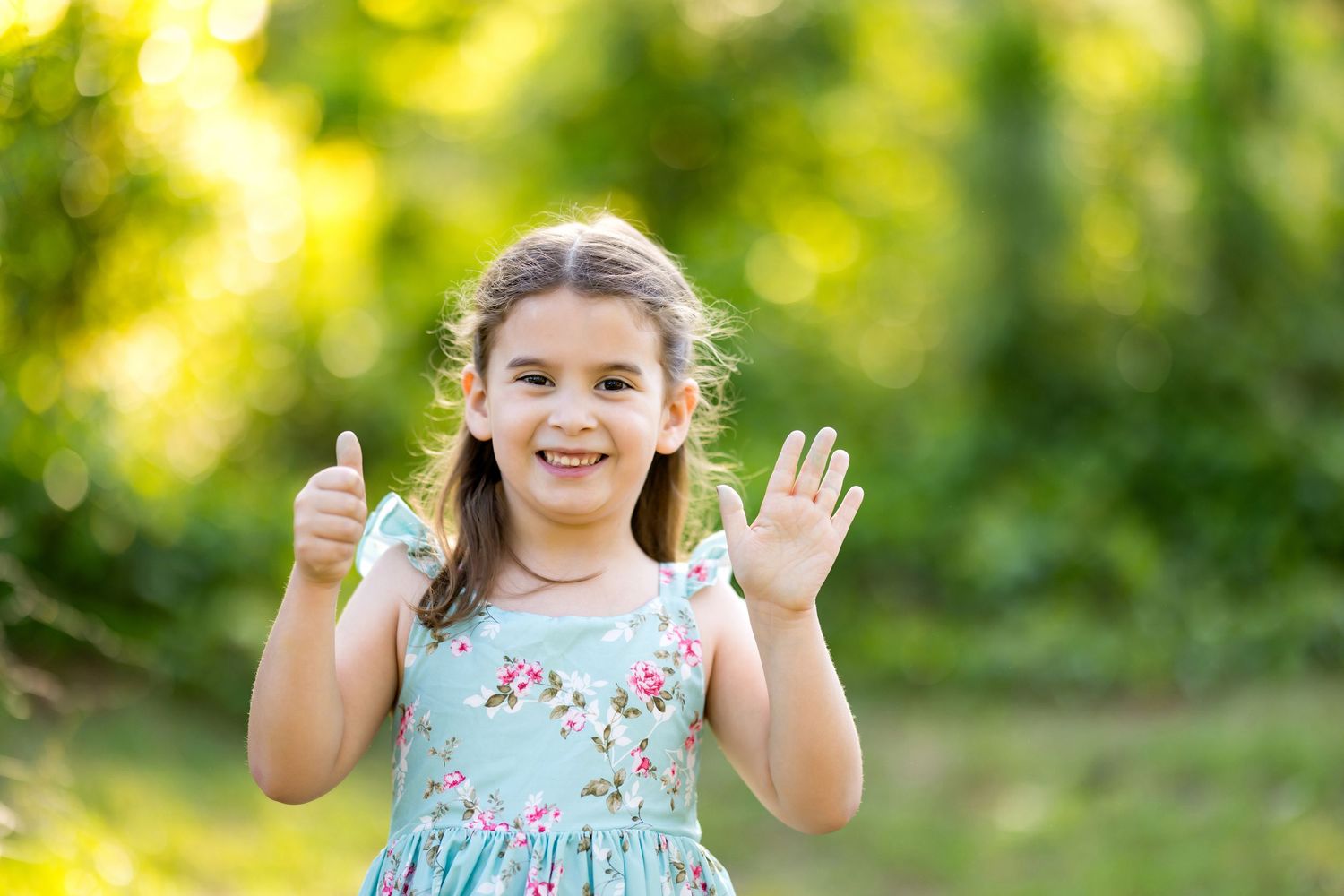 Smiling little girl in floral dress giving thumbs up and waving outdoors in green park setting.