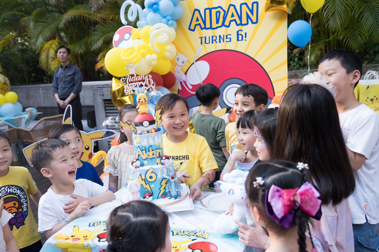 A festive birthday party celebration with yellow and blue balloons, decorations and guests gathered around a table outdoors.