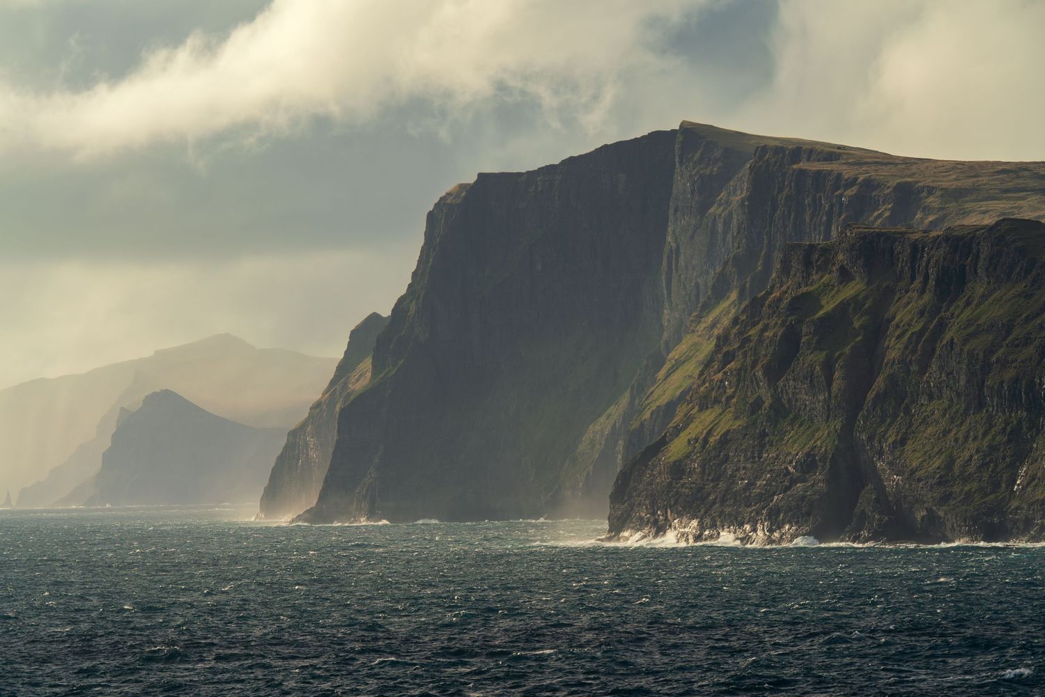 Dramatic coastal cliffs rise above turbulent ocean waters under moody storm clouds in a rugged landscape in the Faroe Islands