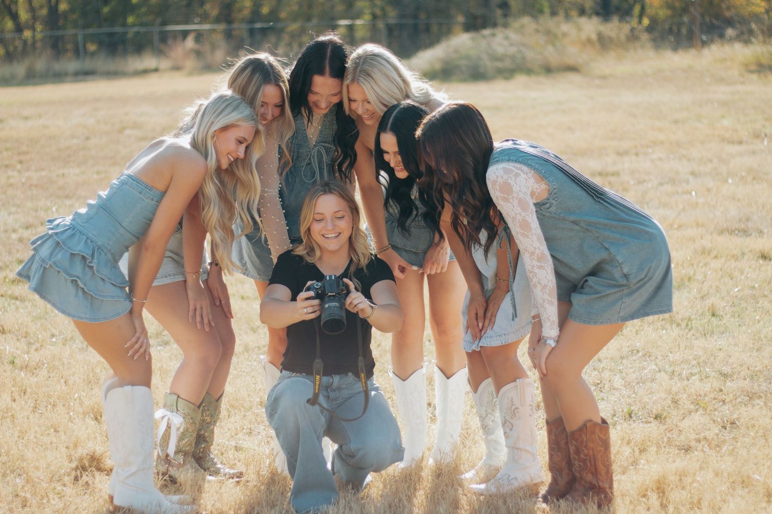 A group of friends in denim and boots huddle together laughing in a sunny outdoor field.
