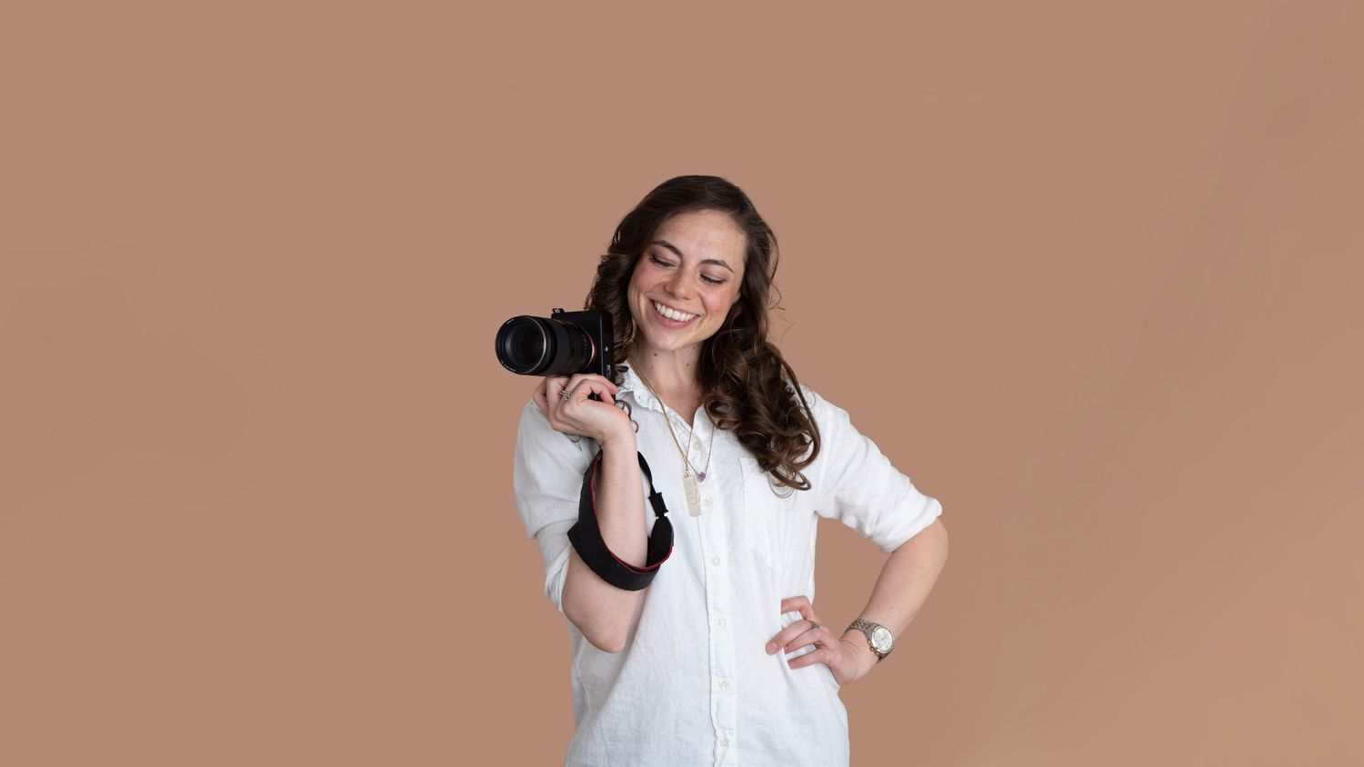Woman in white shirt holding a DSLR camera on her shoulder, posing confidently against a tan background.