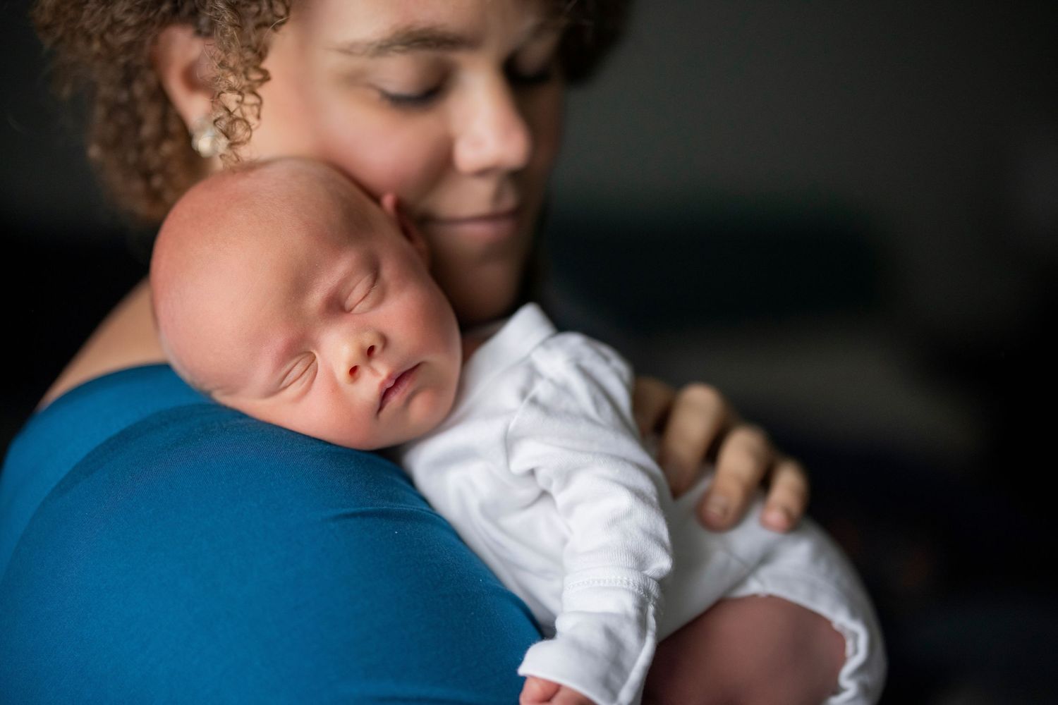 A mother tenderly holds her sleeping newborn baby against her shoulder in a warm, intimate portrait.
