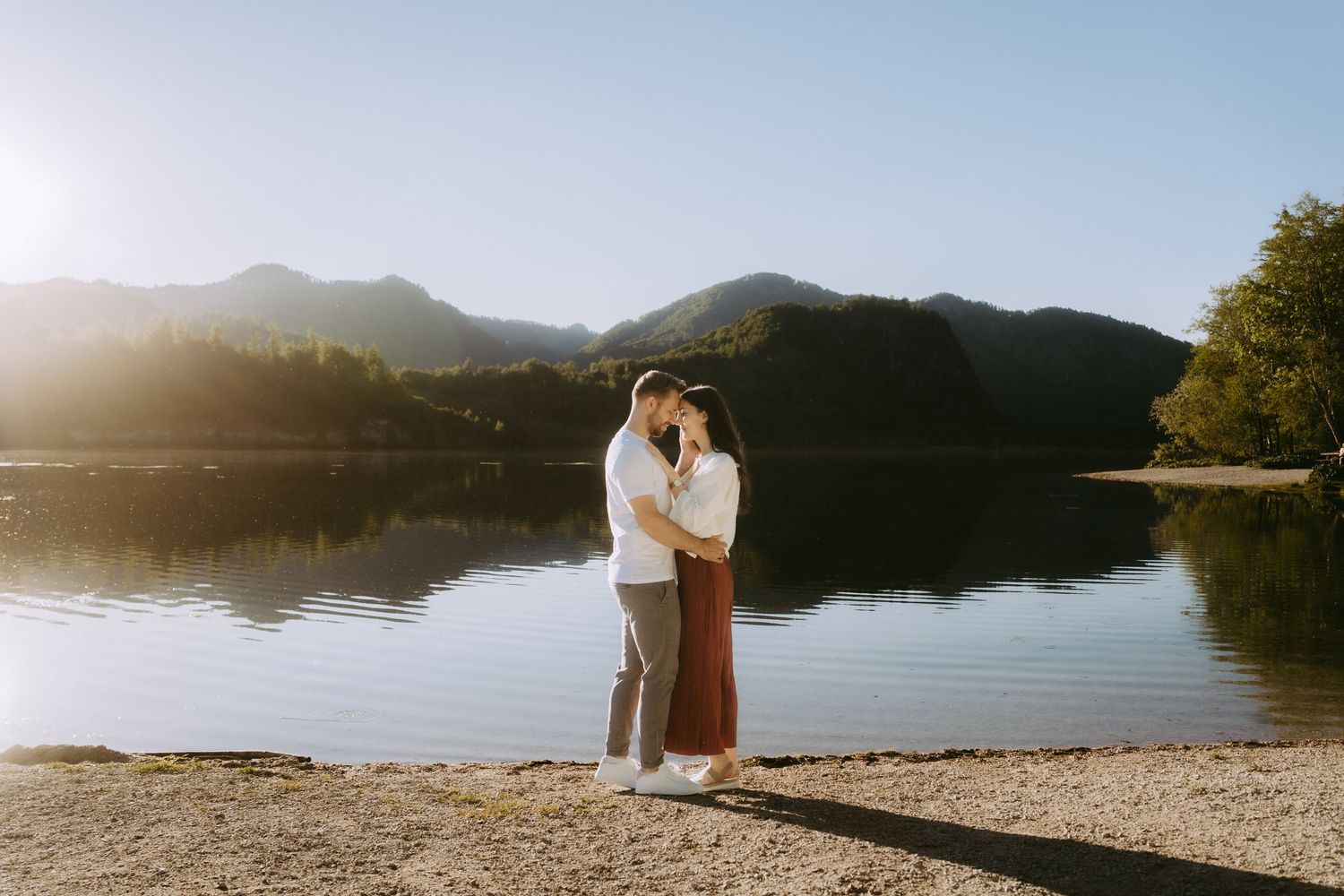 Couple embracing by a serene mountain lake at golden hour with sun flare and reflections in calm water.