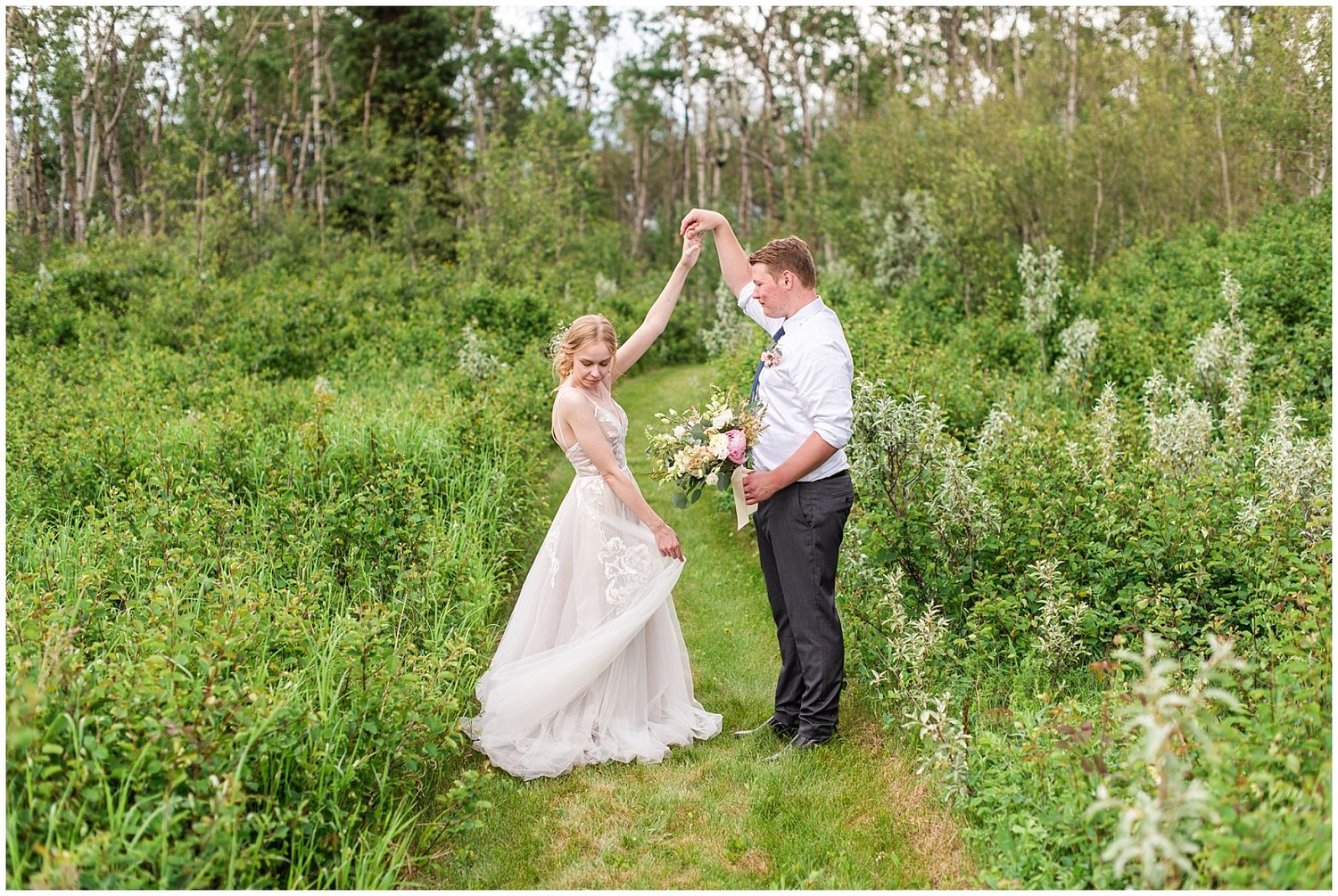 Bride and groom dancing in lush green meadow, groom twirling bride in elegant white gown holding pink bouquet.