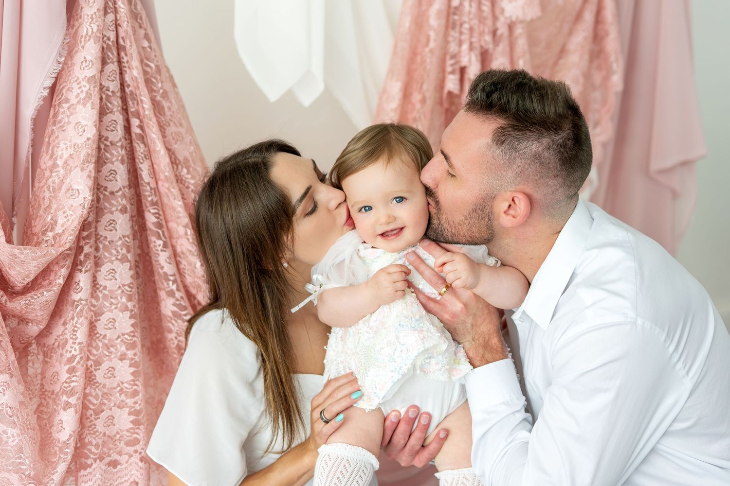 Two parents kiss their smiling baby on the cheeks in a pink lace draped studio.