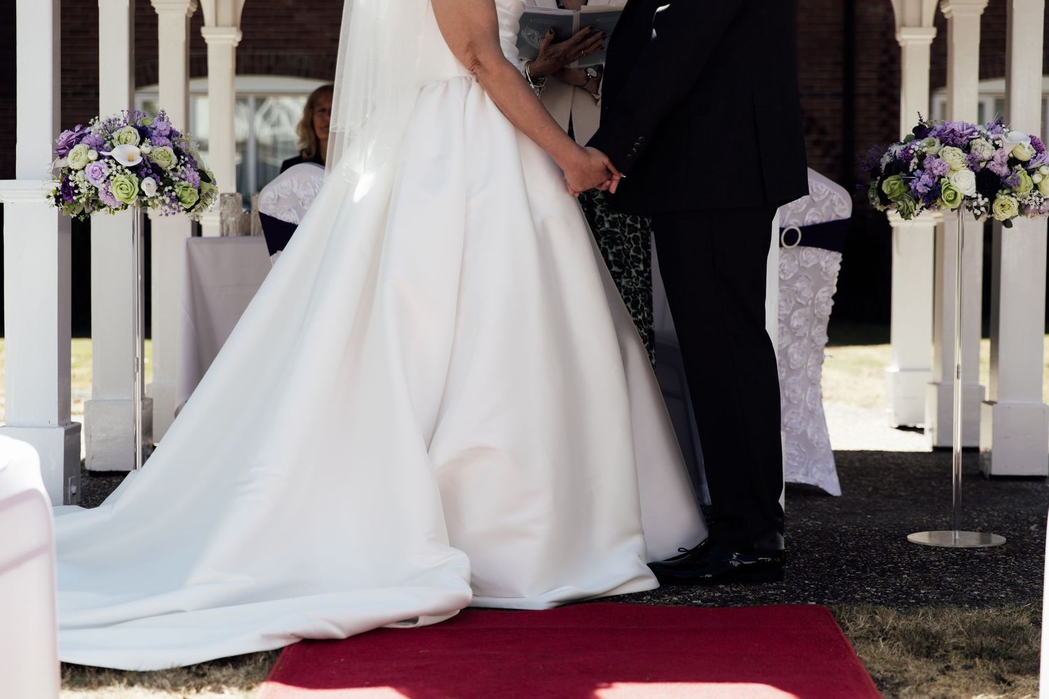 Bride in white gown and groom in black suit exchange vows on red carpet aisle with purple floral arrangements.