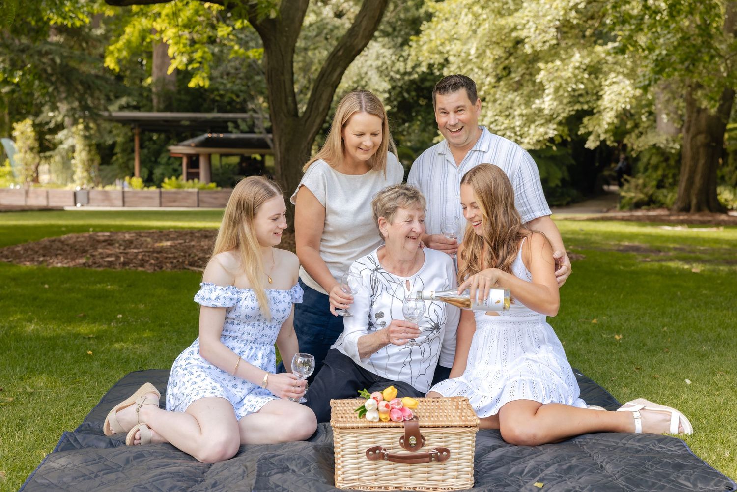 A family group sits together on a blanket in a sunny park setting during a summer picnic gathering.
