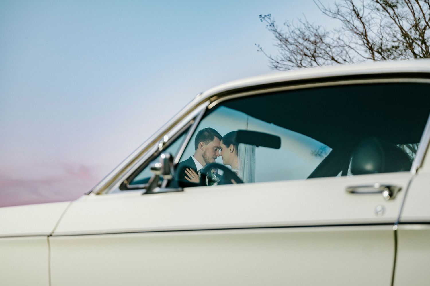 A vintage white car with passengers is seen through the side window against a pastel sky at dusk.