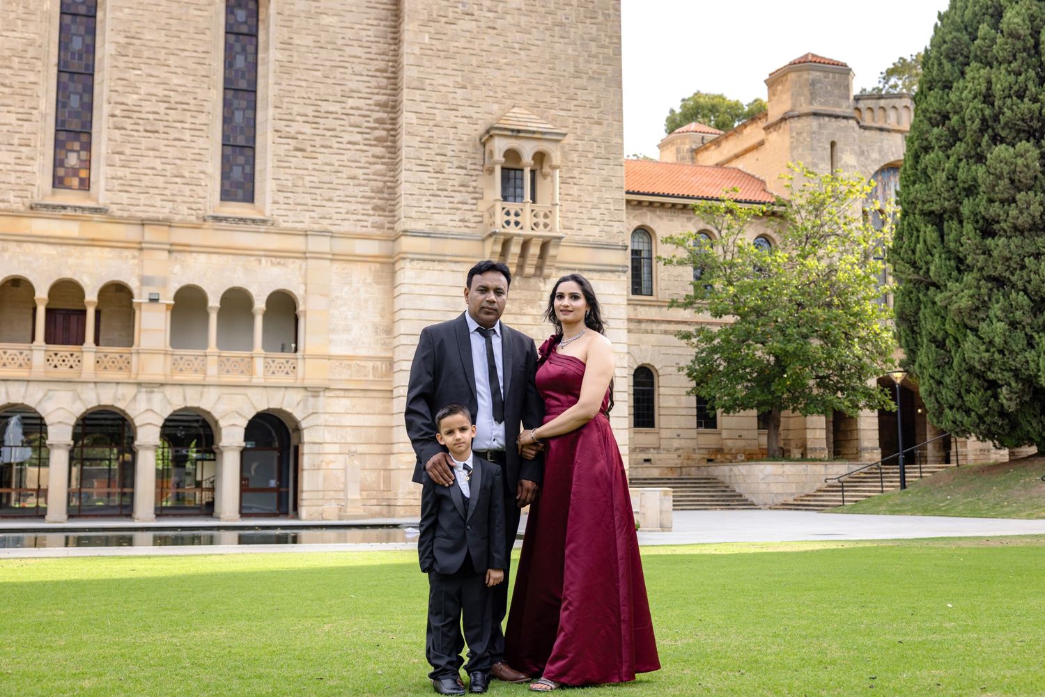 Family of three posing in front of a historic sandstone university building, woman in burgundy gown, man in suit.