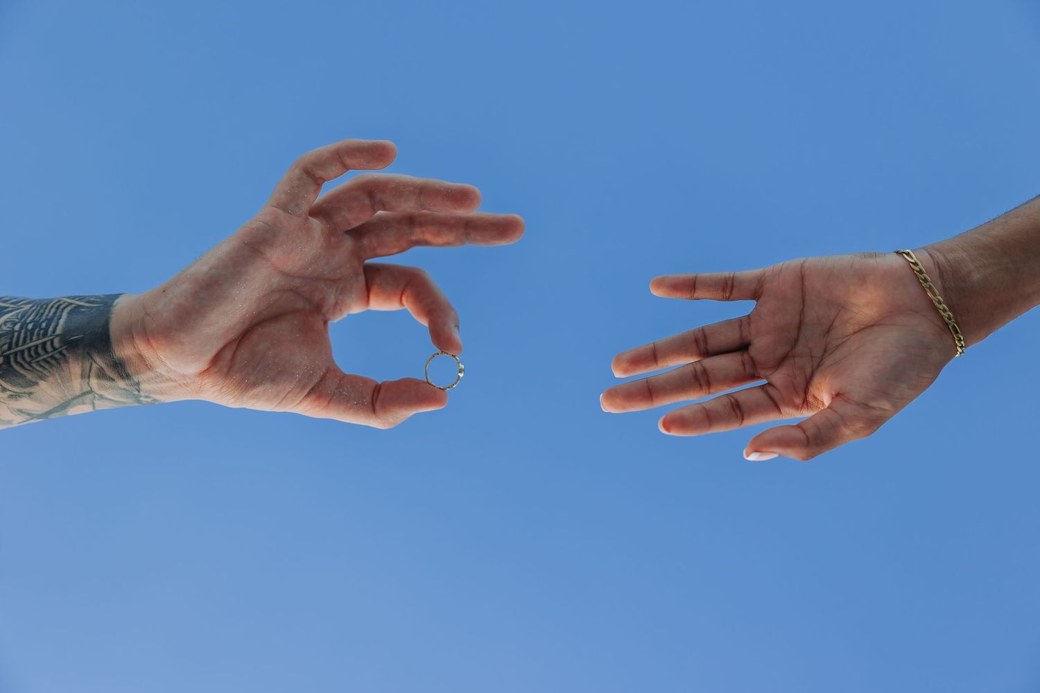 Two hands reaching toward each other against a blue sky, one tattooed holding a small object, the other wearing a gold bracelet.