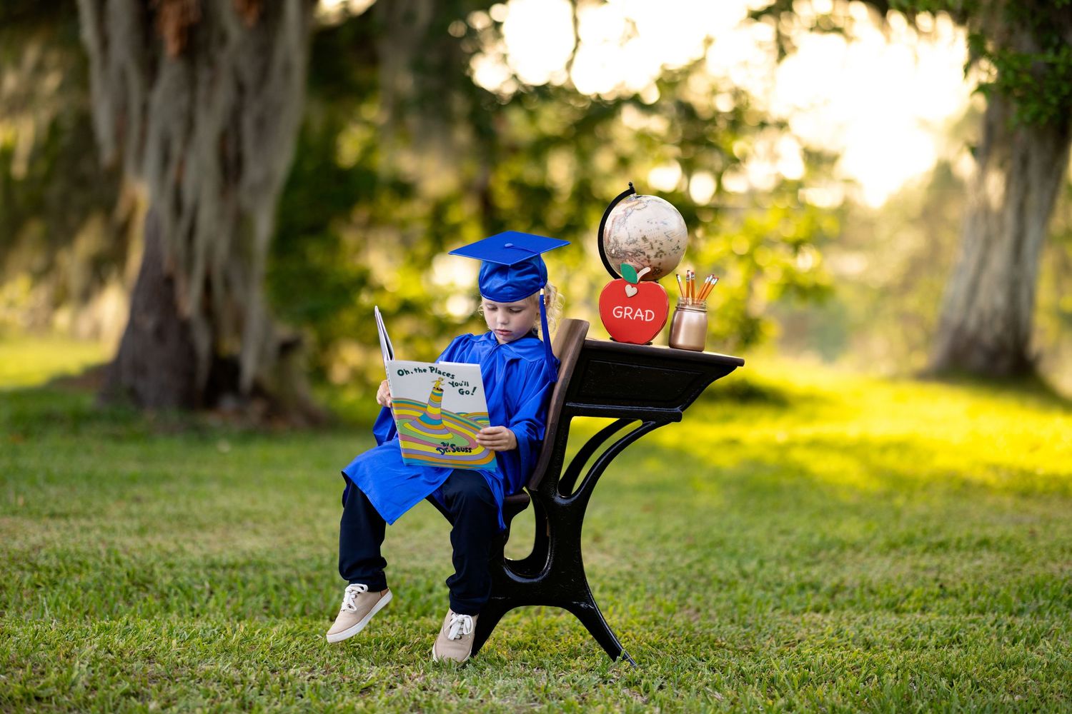Toddler in blue graduation cap and gown sits at school desk reading a book outdoors under large trees.