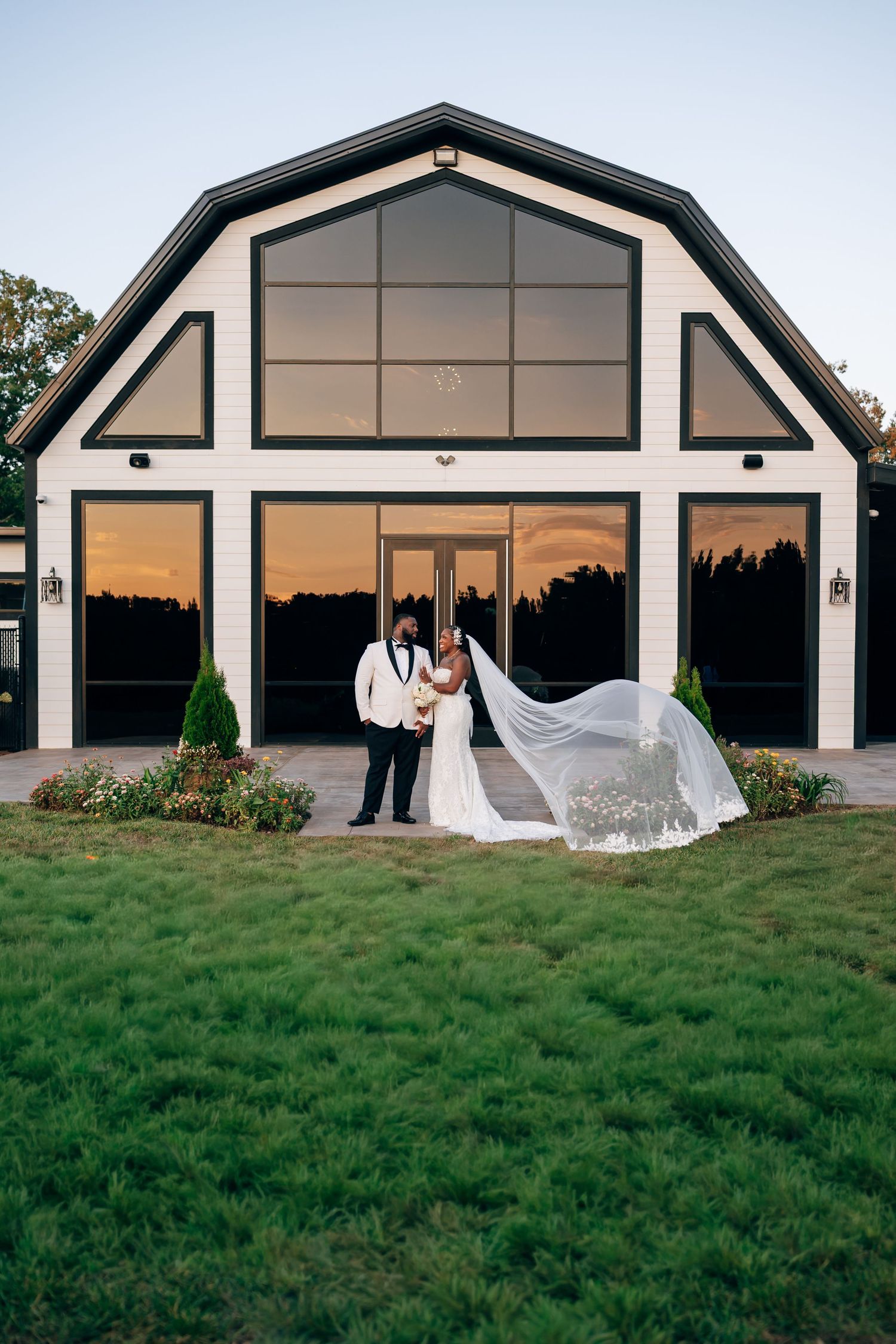 Bride and groom embrace on the lawn outside a modern barn-style wedding venue at sunset as her long veil flows in the breeze.