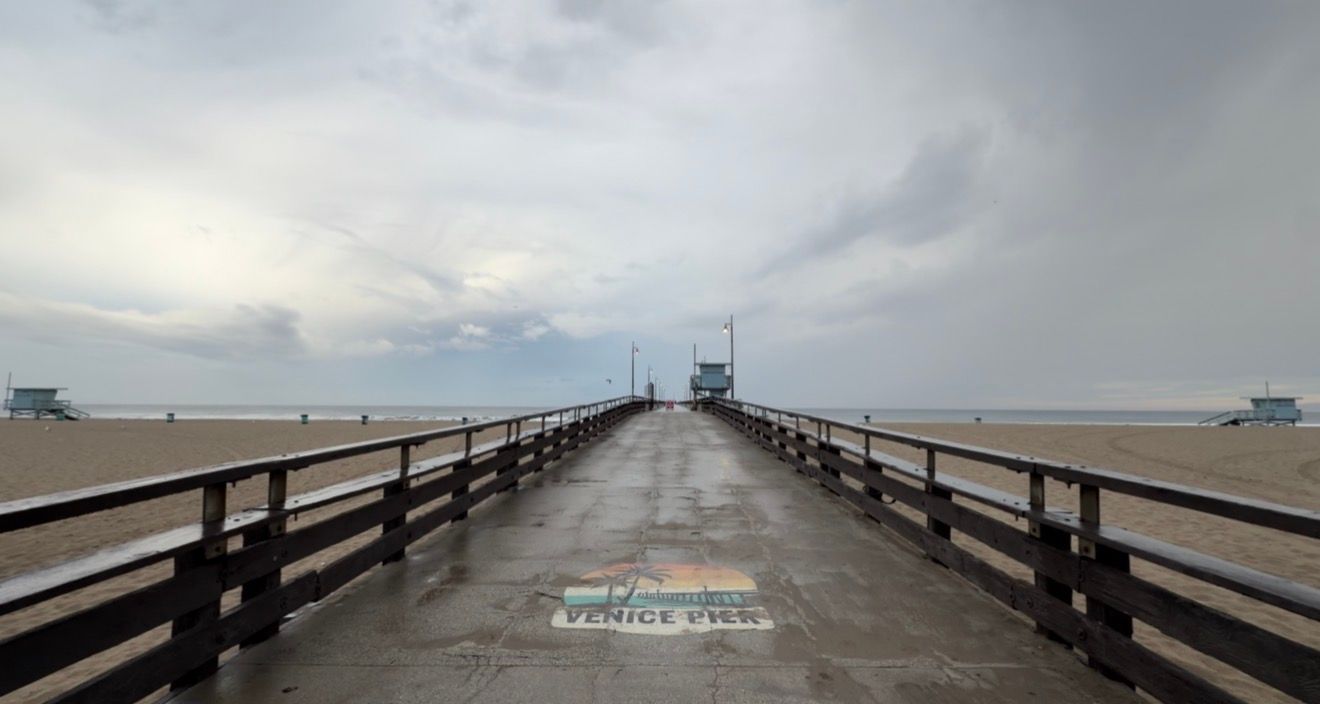 Wet Venice Beach Pier on a cloudy overcast day with calm ocean views and a lifeguard tower in the distance.
