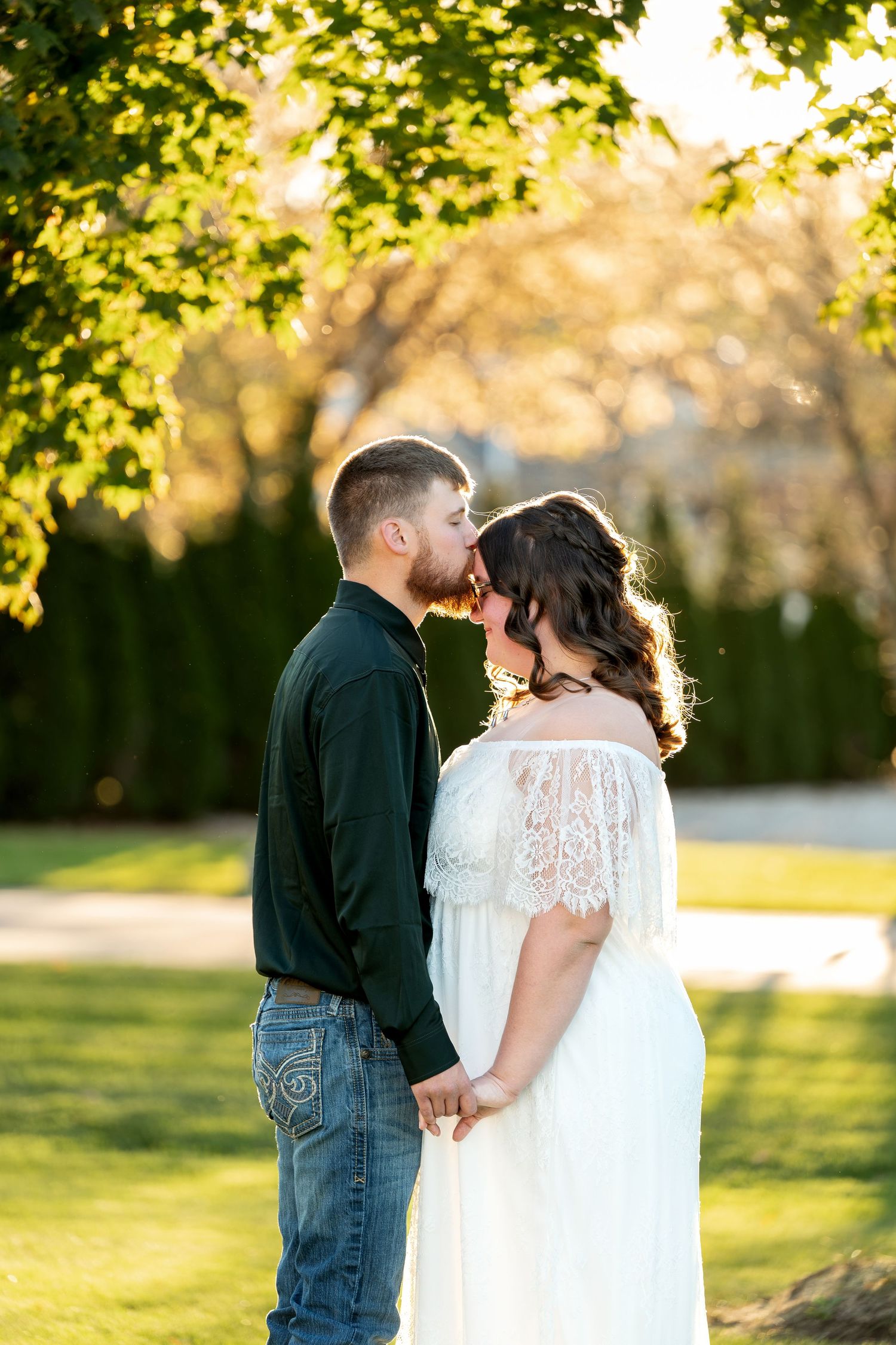 A couple shares a romantic kiss outdoors, holding hands in golden sunlight, she in a white lace gown, he in a dark shirt.