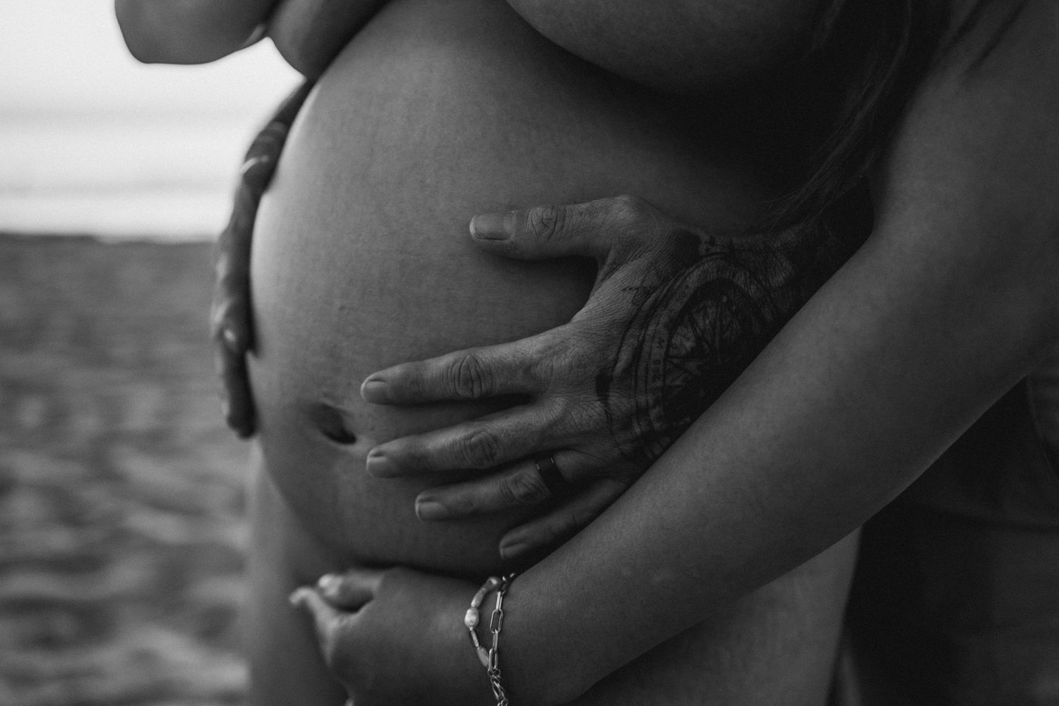 Black and white photo of a pregnant woman cradling her baby bump on a beach with a partner's hands.