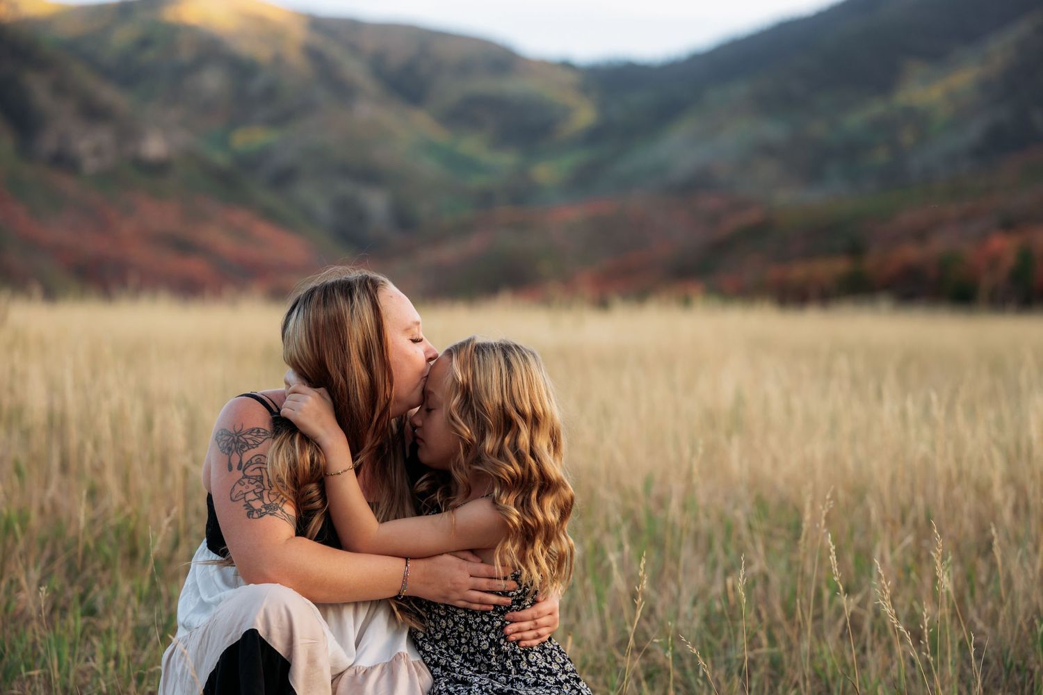 Two women sharing a warm embrace and kiss in a golden meadow with colorful autumn hills in the background.