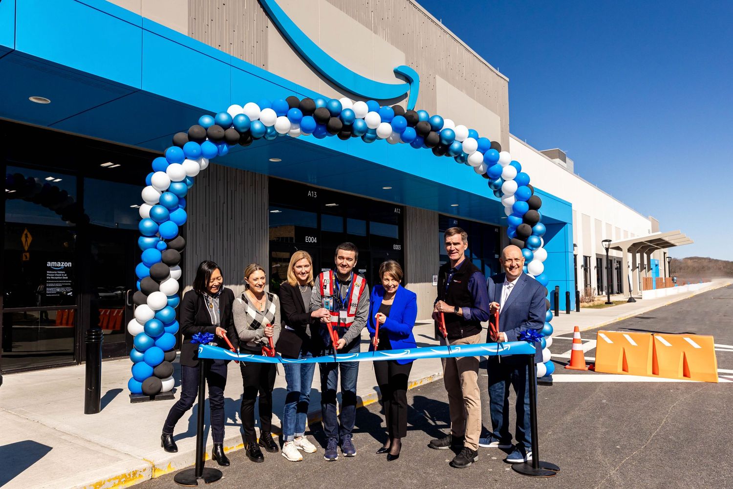 Group gathers for ribbon cutting ceremony under blue and white balloon arch at store opening.