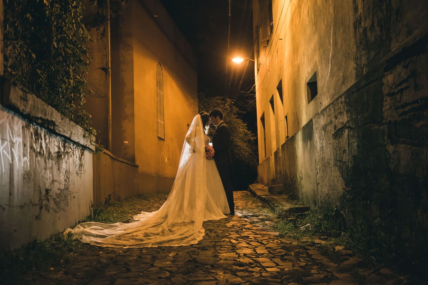 Couple in wedding attire pose in dimly lit cobblestone alley at night.