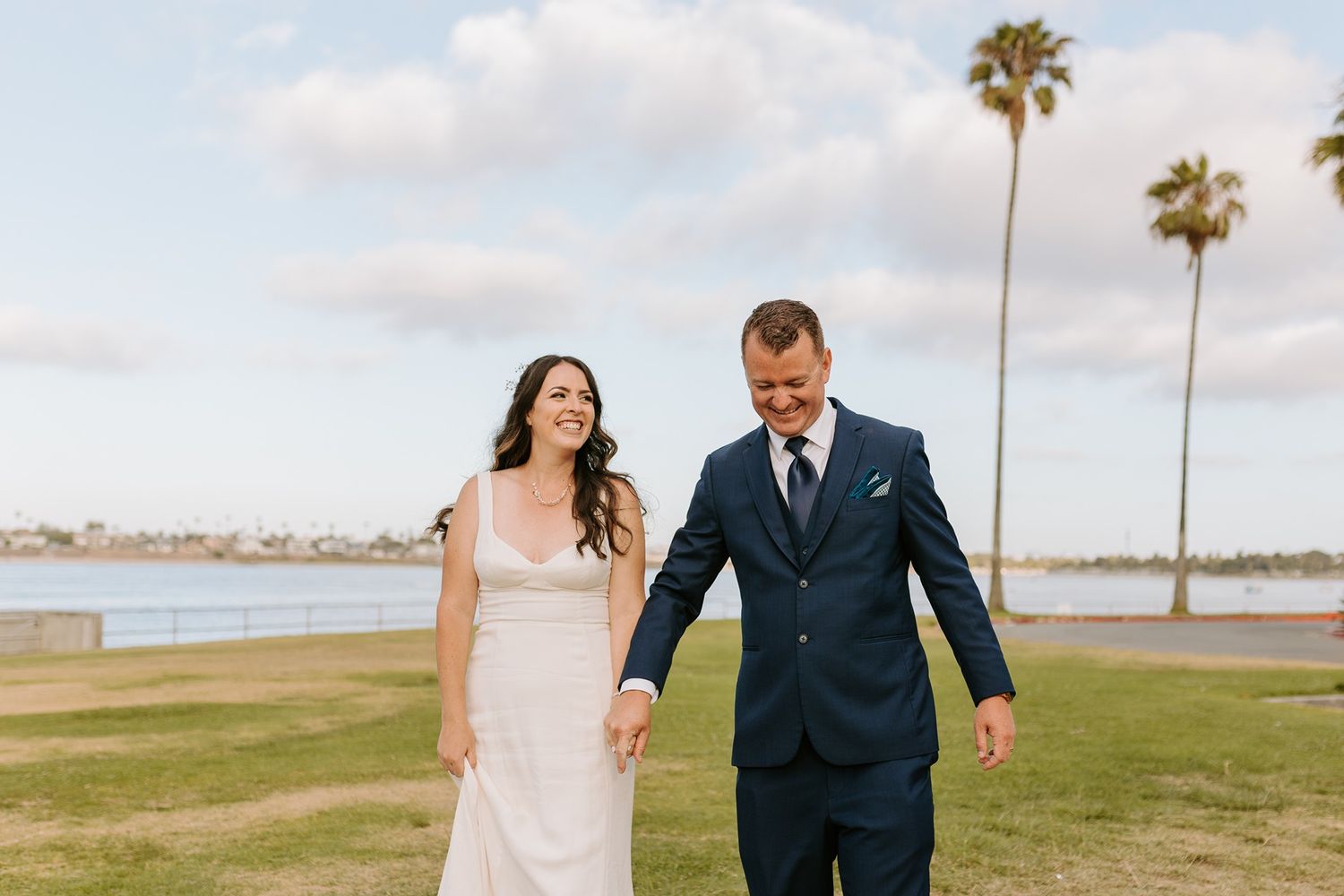 Wedding couple walks hand in hand on a grassy waterfront with palm trees in the background at Tower Beach Club.