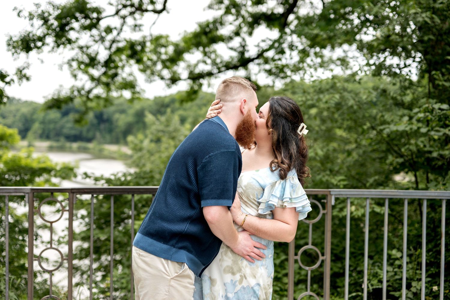 Couple sharing a romantic kiss on a scenic overlook bridge surrounded by lush green trees and a river below.