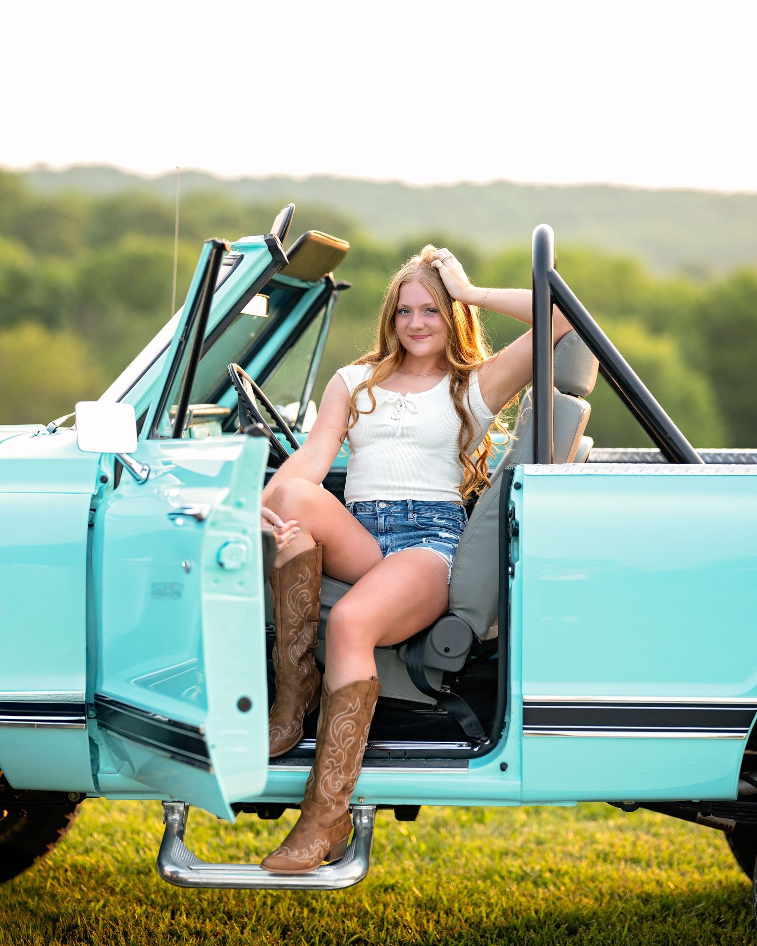Young woman in denim shorts and cowboy boots posing in a vintage teal convertible car outdoors. Kansas City senior portrait session - natura