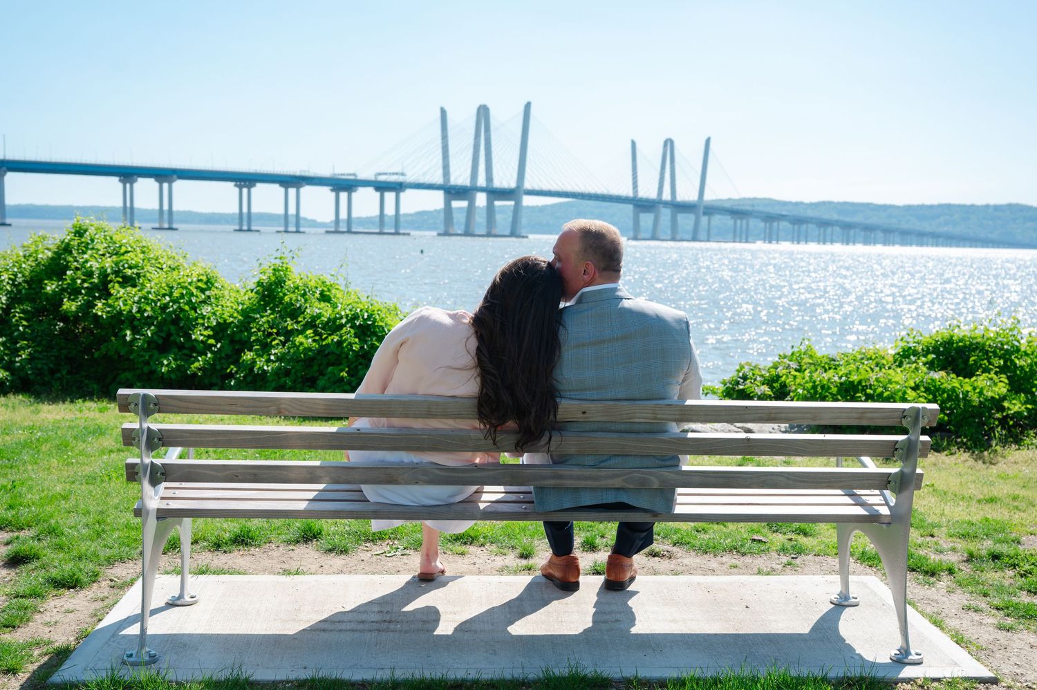 Couple sitting on a white bench kissing with a scenic river bridge view on a sunny day.