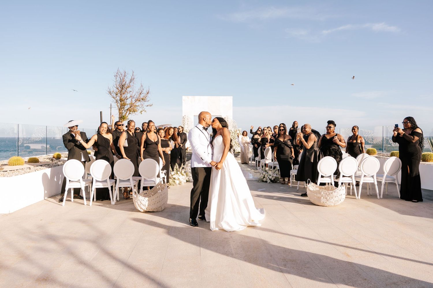 Outdoor wedding ceremony on a rooftop with white floral arrangements, a couple exchanging vows, and birds flying overhead.