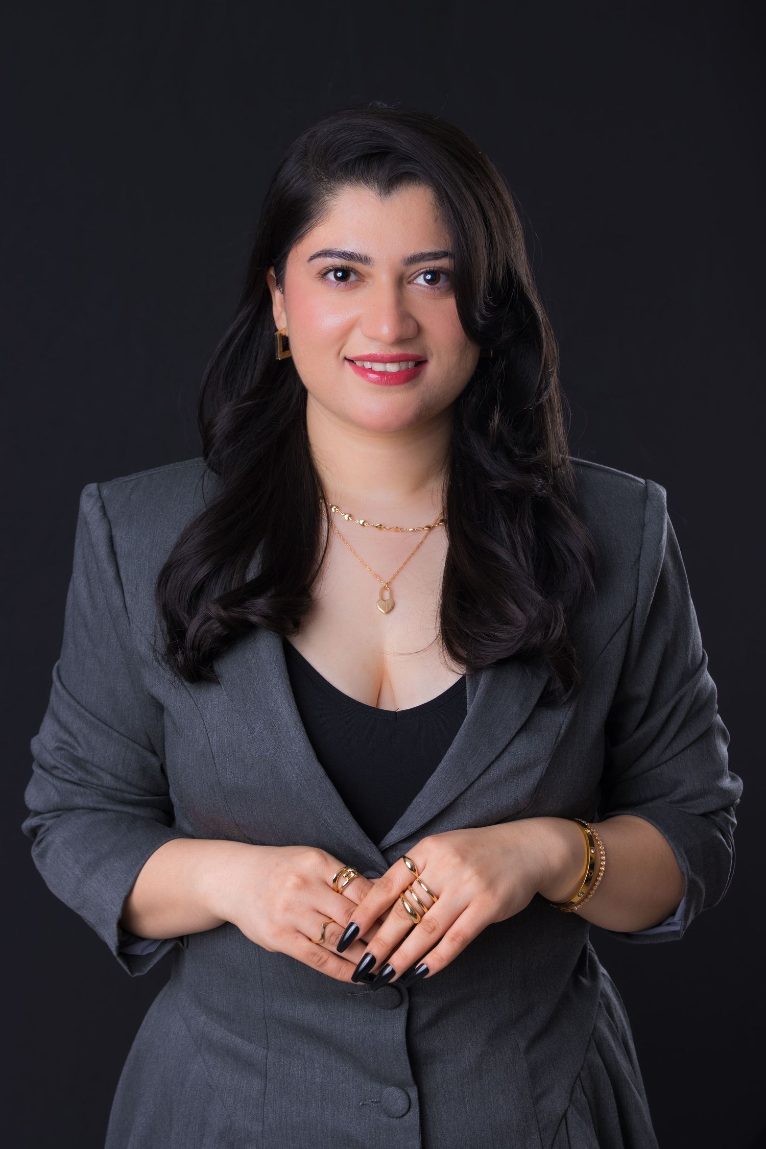 Professional woman in gray blazer with gold jewelry smiling confidently against dark background.
