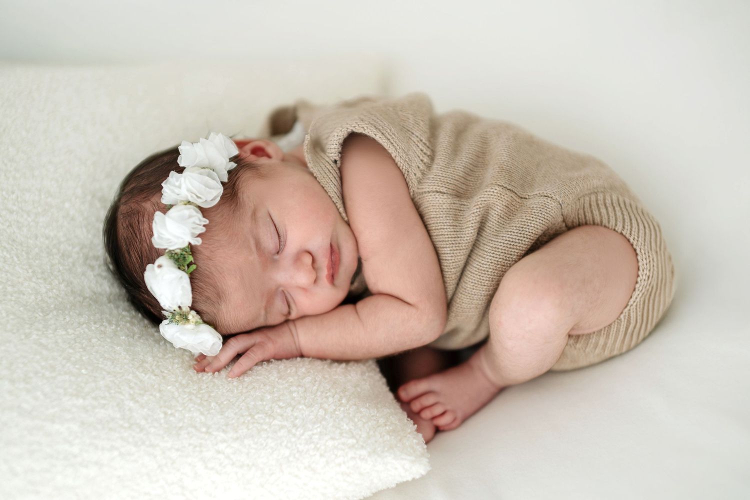 A sleeping newborn in a beige knit outfit and white floral headband rests on a soft white blanket.