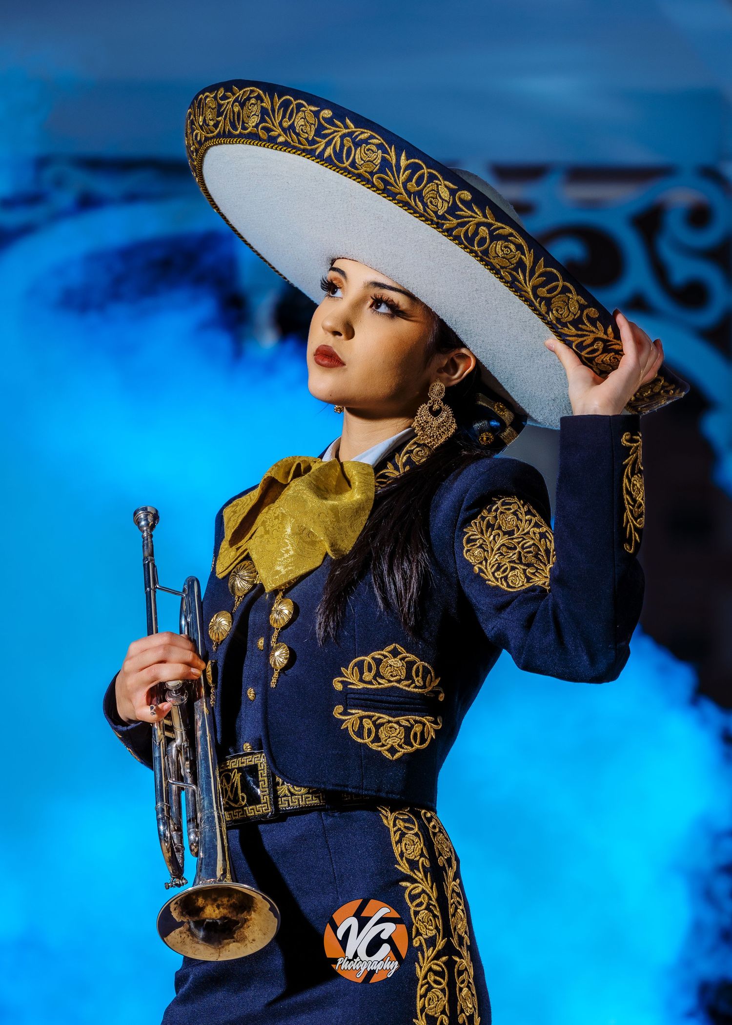 A woman in a mariachi outfit holds a trumpet while posing in a navy and gold charro suit with a large sombrero.