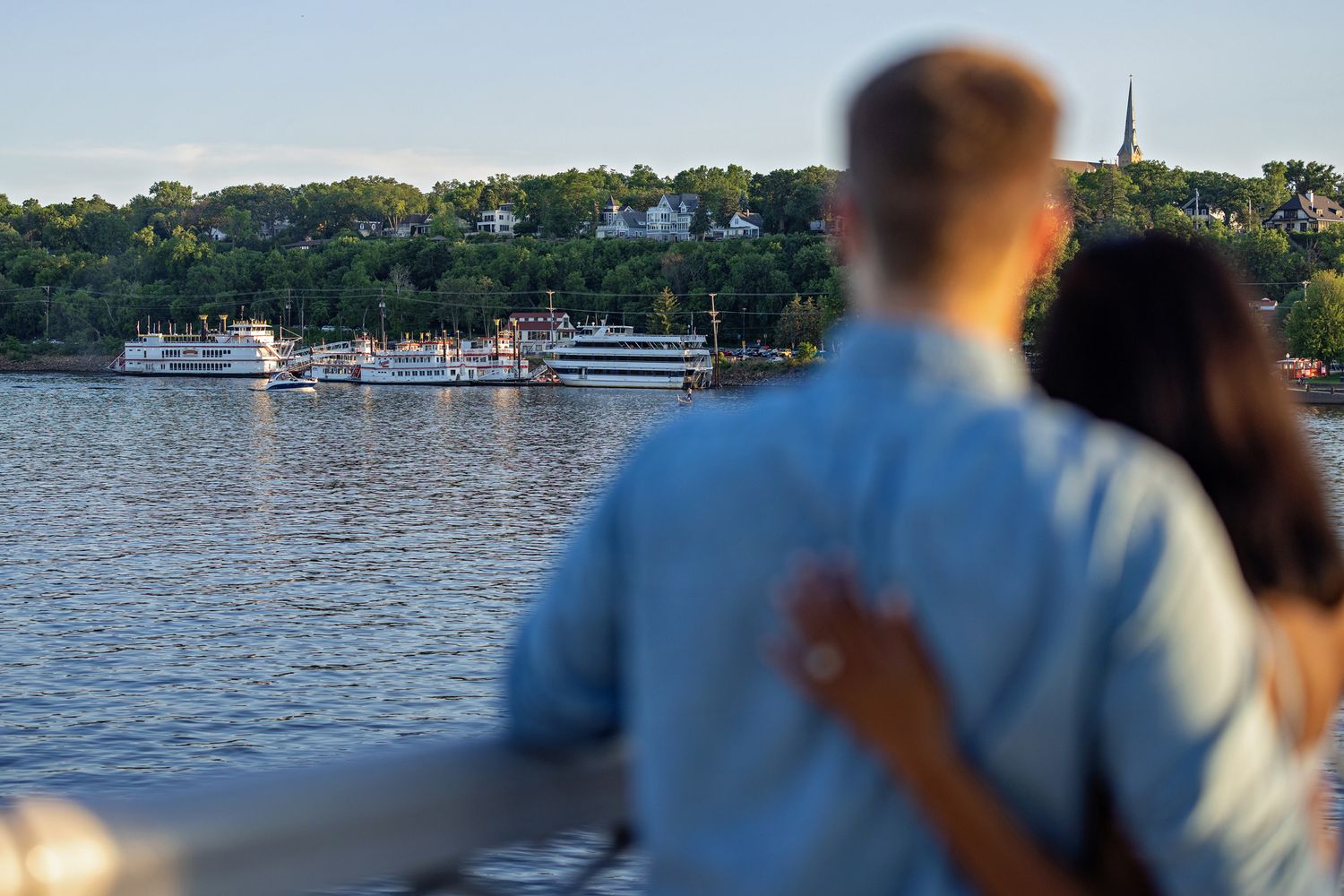 Couple embracing on a lift bridge overlooking the St. Croix River with a ferry boat and tree-lined Stillwater, MN shore in the background.