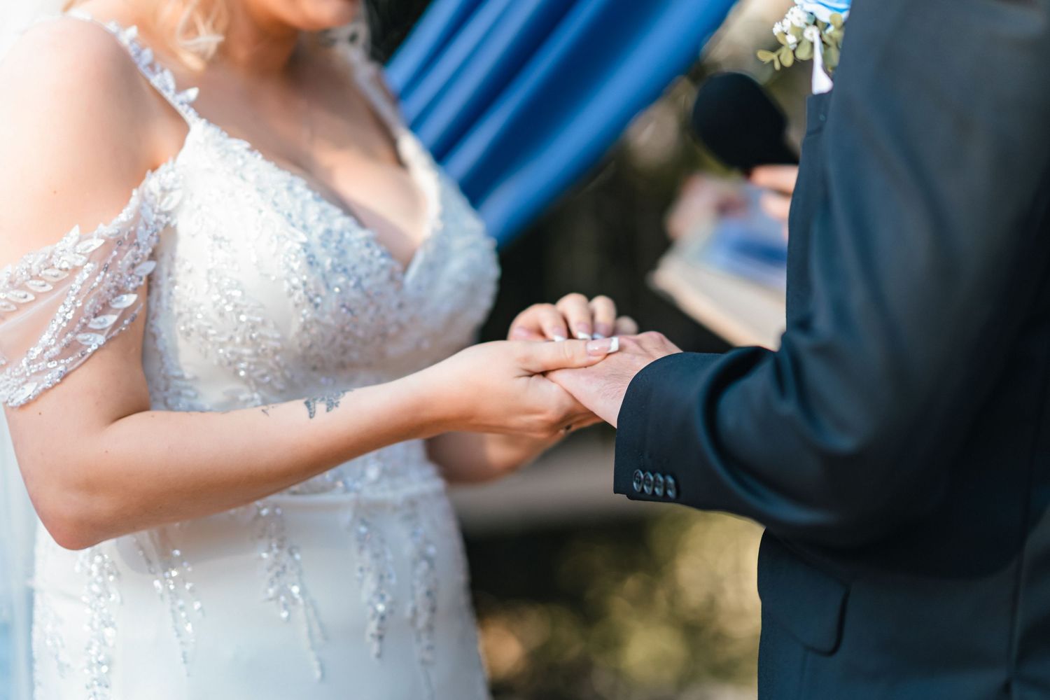 Bride and groom exchanging rings during outdoor wedding ceremony, bride wearing elegant beaded white gown.
