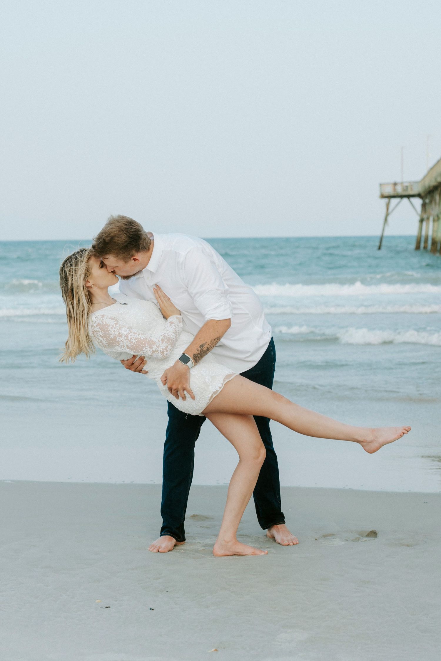 A couple shares a romantic kiss on the beach, as the groom lifts his bride in a white lace dress by the ocean.