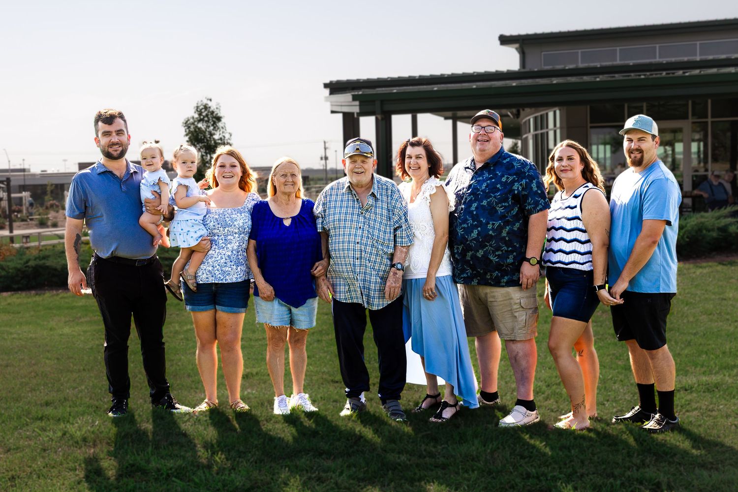 A large family group of nine people smiling outdoors on green grass near a modern building on a sunny day.