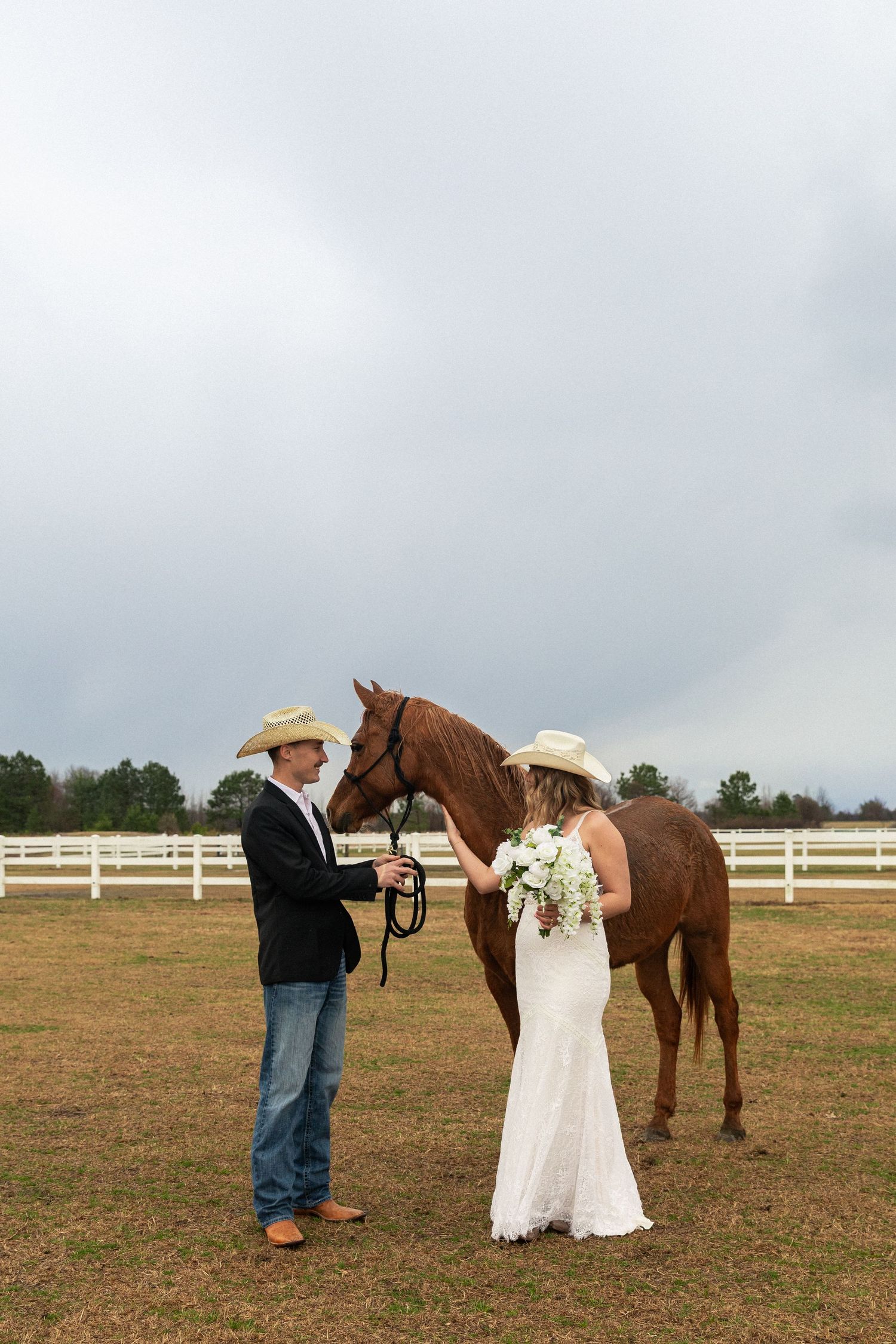 Bride and groom in cowboy hats stand with a chestnut horse at a white-fenced ranch on their rainy wedding day.