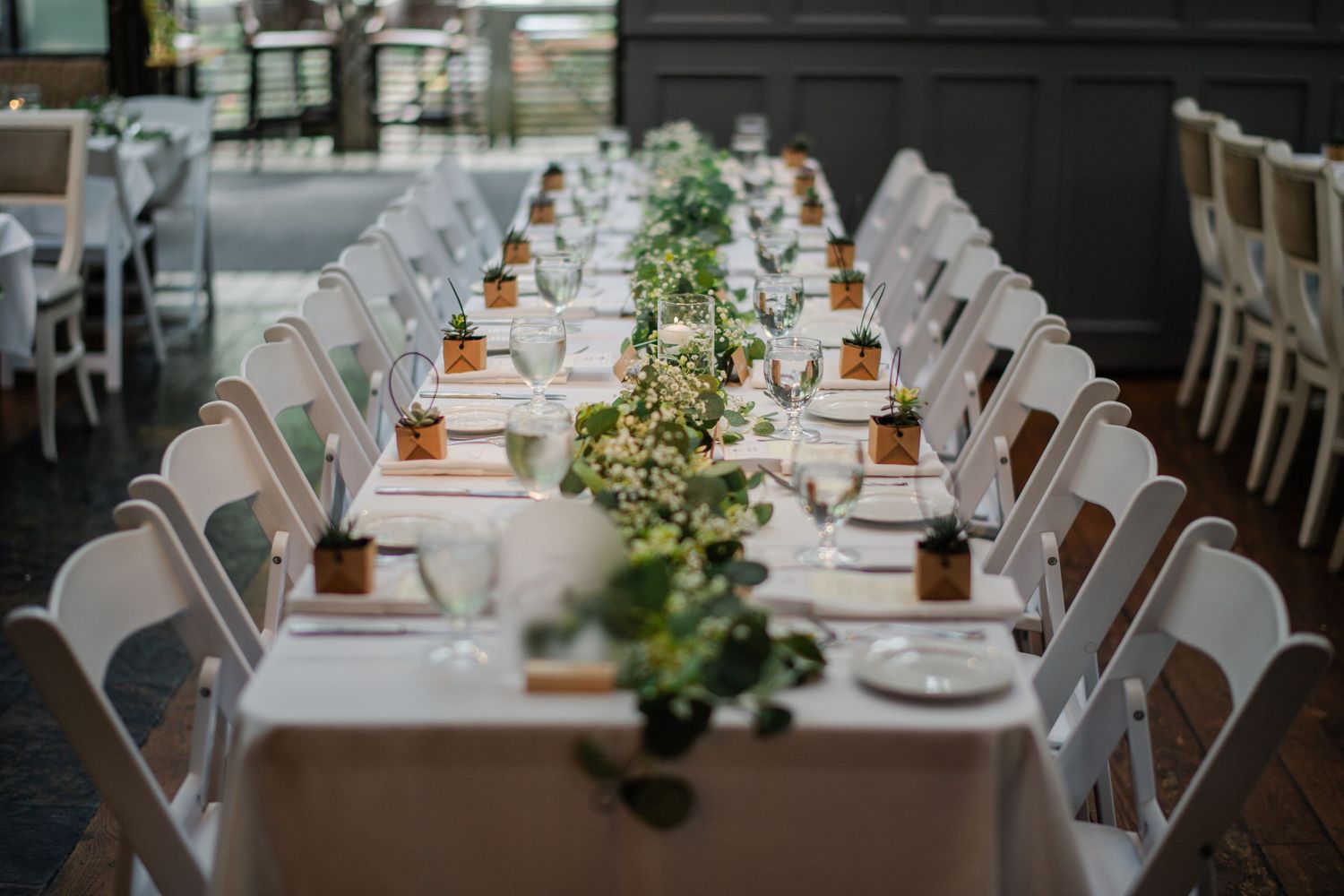 Elegant wedding reception venue with white-draped tables, greenery centerpieces, string lights, and large windows overlooking lush trees.