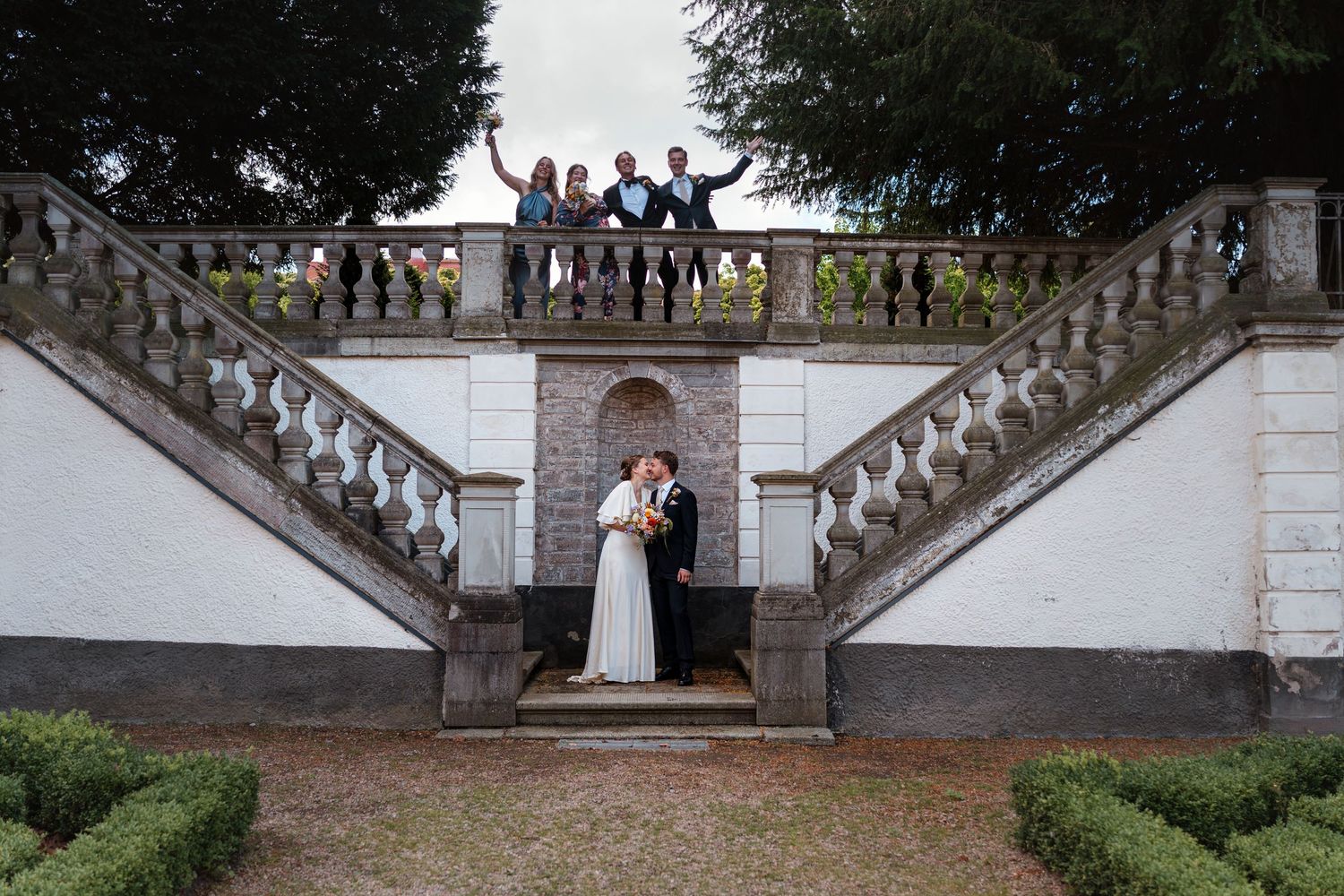 Wedding couple kissing under grand staircase with guests celebrating on balcony above at elegant outdoor venue.
