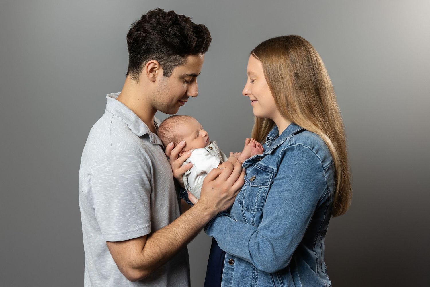 Young couple smiling and holding a sleeping newborn baby against a gray studio background.