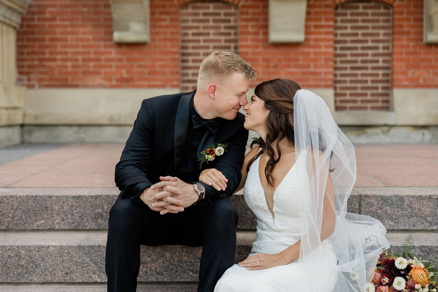 Romantic wedding couple shares tender moments in front of brick building with autumn floral bouquet.