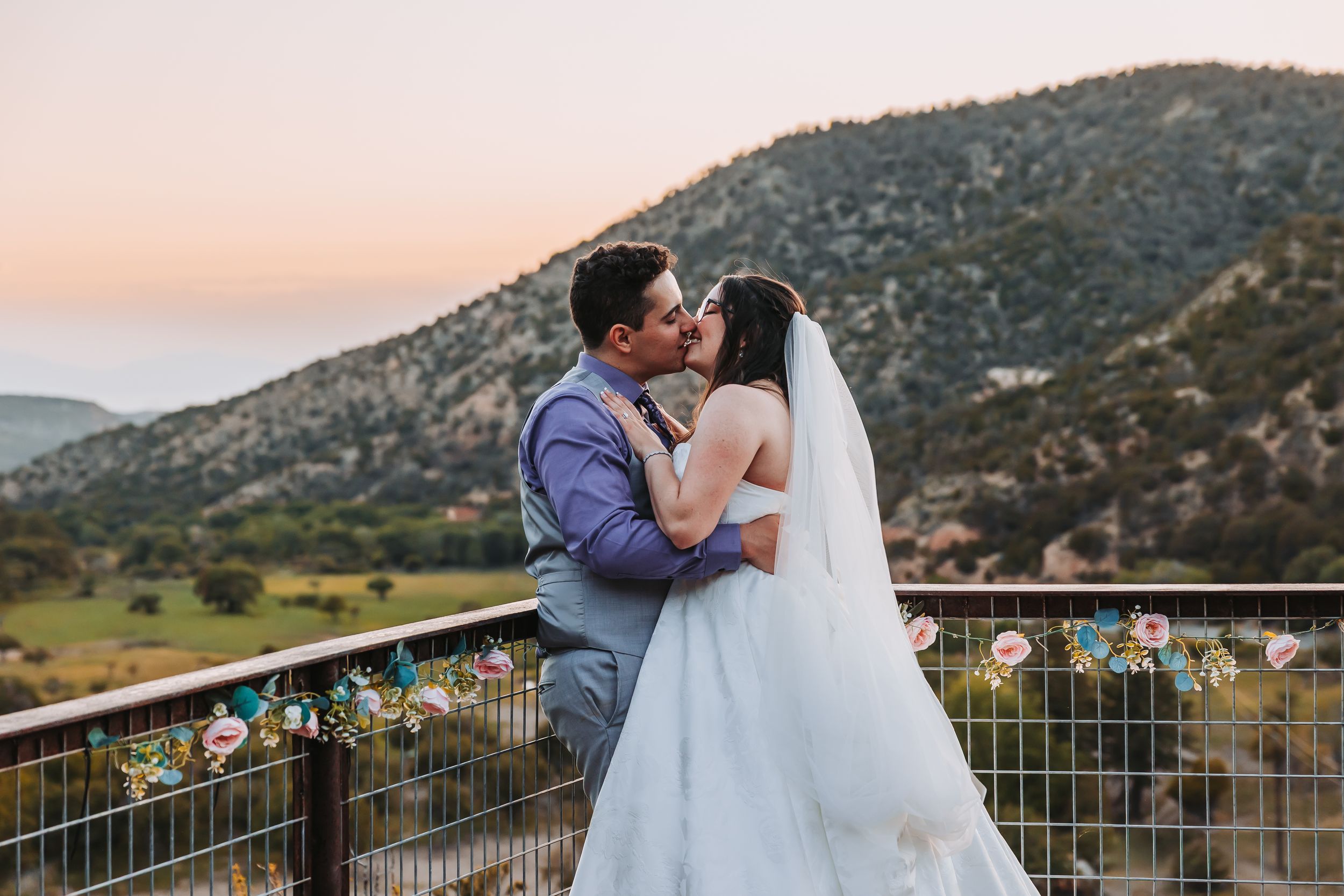 Bride and groom share a romantic kiss on a decorated balcony with scenic mountain views at sunset.