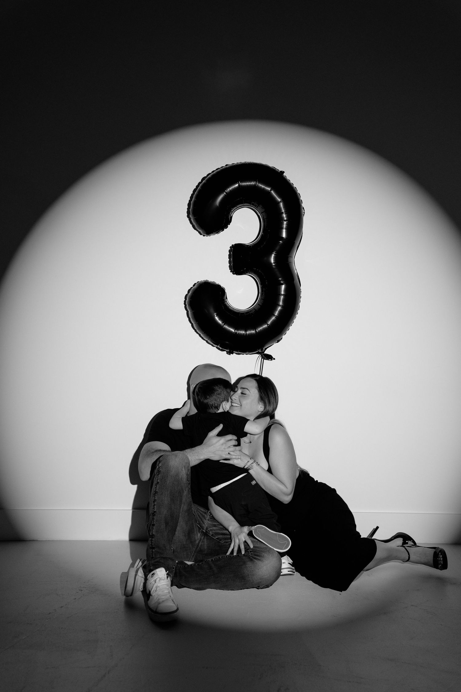 Black and white photo of a couple kissing on the floor beneath a large number 3 balloon in a spotlight.