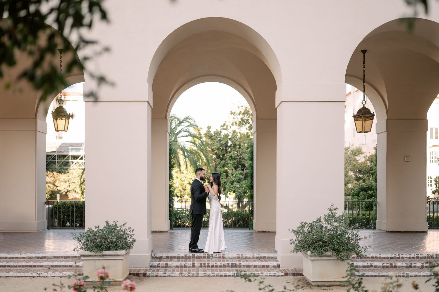 Bride and groom share a romantic moment under elegant arched colonnade surrounded by lush greenery at outdoor venue.